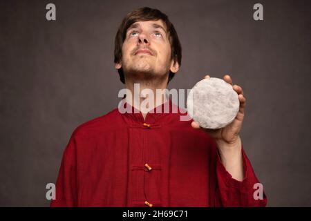 Young handsome tall slim white man with brown hair looking up holding pu erh tea cake in red shirt on grey background Stock Photo