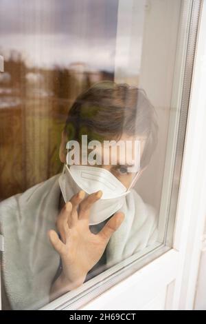 Young handsome tall slim white man with brown hair facing camera looking out the window wearing face mask in white blanket Stock Photo