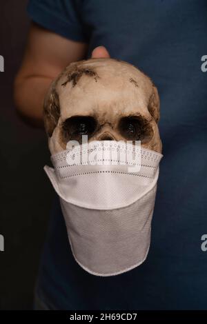 White man holding human skull that wears face mask in blue shirt on grey background Stock Photo