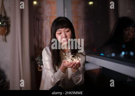 A young, beautiful Korean Asian woman holds a lighted festive garland in her cozy house with dreams of positive emotions in the evening on the window Stock Photo