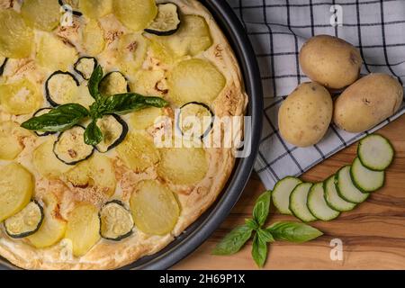 A pan of quiche with potatoes and sliced zucchini on a wooden cutting board with whole potatoes beside it Stock Photo