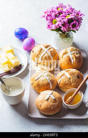 Traditional Easter cross buns with raisins, butter and honey on gray background with flowers and colored eggs. Stock Photo