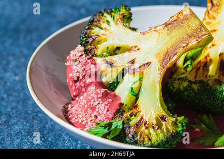 Grilled broccoli steak with pink beetroot sauce and hemp seeds in white bowl, blue background, close-up. Healthy vegan food concept. Stock Photo