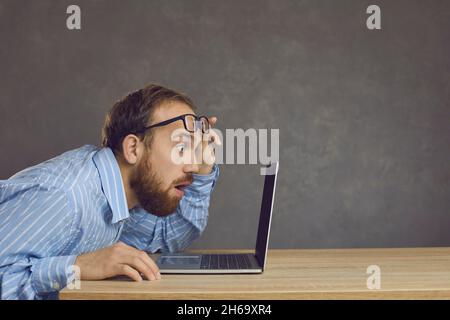 Side view of a man with a surprised expression sitting at a table and staring at a laptop screen. Stock Photo