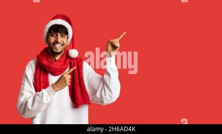 Positive middle eastern young man in red Santa hat, scarf and white pullover pointing at copy space for Christmas or New Year advertisement over red s Stock Photo