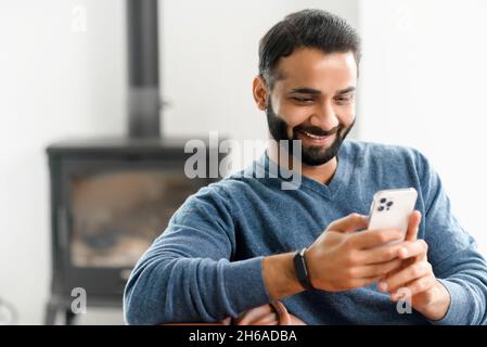 Smiling Indian guy wearing casual outfit messaging, texting on the smartphone.Multiracial bearded man sits on the modern sofa and using mobile app, scrolling feed news, websurfing, playing game Stock Photo