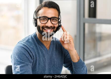 Headshot of young Indian male wearing handsfree headset and stylish glasses, in casual wear in the office, taking call and talking with customers or colleagues. Call center, help line, support service Stock Photo
