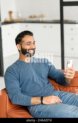 Video call concept. Hilarious dark-haired bearded Indian man in casual wear using smartphone for virtual meeting, enjoys online chatting sitting on the sofa at home, guy watching movie and laughing Stock Photo