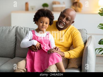 Smiling little african american girl sitting on knees of elderly grandfather on couch in living room interior, copy space. Happy together indoor at fr Stock Photo