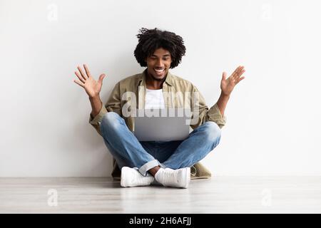 Online Success. Surprised Young Black Guy Looking At Laptop Computer Screen With Excitement, Happy African American Man Raising Hands And Exclaiming W Stock Photo