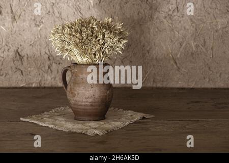 Abstract rural composition of clay vase with a bouquet of dried cereals standing on gray linen tablecloth and old, grunge and aged wooden desk in fron Stock Photo