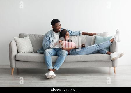 Leisure Time. Smiling cheerful millennial black couple using pc at home, browsing internet together, lady lying on guy's lap, sitting on comfy sofa in Stock Photo