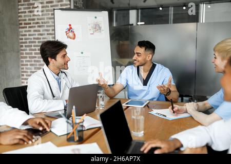 Cheerful multiracial doctors men and women in uniforms sitting aroung table at clinic, having conversation, using modern gadgets, sharing professional Stock Photo