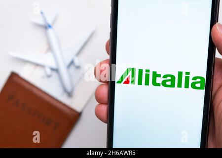 Alitalia Airline application on the smartphone screen mans hand. A white toy airplane and a passport are lying on a table with a light surface.November 2021, San Francisco, USA Stock Photo