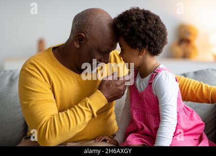 Happy elderly african american man and little girl touches foreheads and enjoying time together at home interior. Love, tenderness, family and relatio Stock Photo