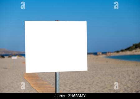 Sign white blank on post, sandy beach background. Outdoors empty signboard billboard signage label on metal pole. Sunny day summer holiday advertise t Stock Photo