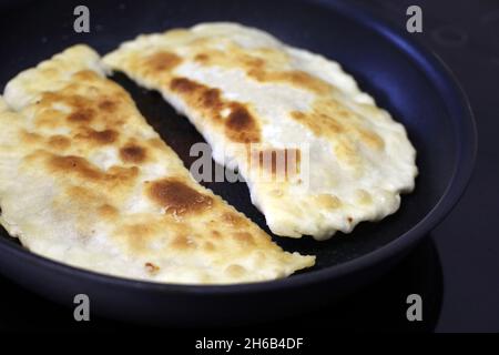 Frying chebureks on electric induction stove, traditional eastern dish. Homemade tortillas with meat on black pan fried in oil Stock Photo