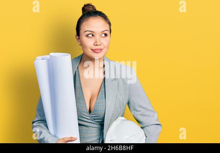 Young hispanic girl holding safety helmet and blueprints smiling looking to the side and staring away thinking. Stock Photo