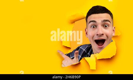 Amazed shocked emotion happy young european man with open mouth look through hole in yellow paper and point finger on empty space, collage, panorama. Stock Photo