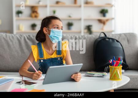 Self-Isolation, Staying At Home. Sad upset adolescent student wearing surgical medical face mask, sick kid studying in living room using her tablet, l Stock Photo