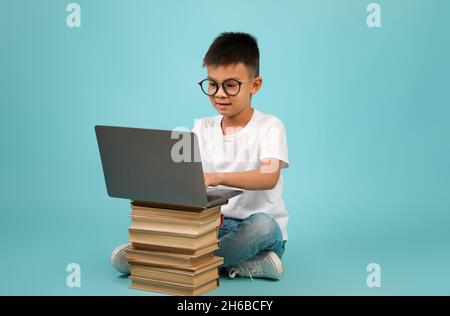 Distant Learning Concept. Cute Asian Schoolboy In Eyeglasses Using Laptop Placed On Book Stack, Smart Korean Male Kid Study Online With Computer, Sitt Stock Photo