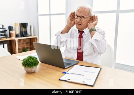 Senior man working at the office using computer laptop trying to hear both hands on ear gesture, curious for gossip. hearing problem, deaf Stock Photo