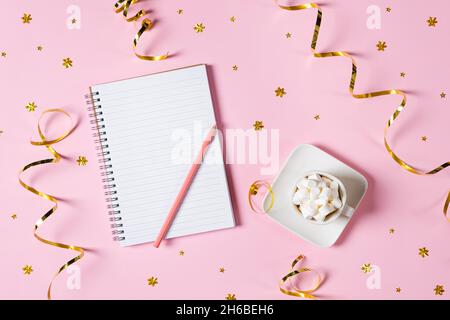 New year, Christmas or holiday wish list concept. Notepad, golden tinsel, and a cup of hot chocolate with marshmallows isolated on pink background Stock Photo