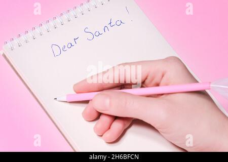 Dear Santa letter concept. Hand holding pink pen decorated with feather and writing in Notepad letter to Santa Claus on pink background. Stock Photo