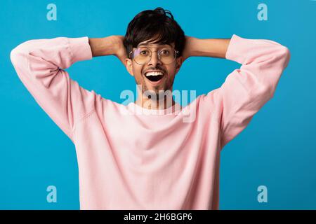 Amazed young indian man in pink wearing glasses looking at camera and gesturing, holding hands behind his head, looking at something exciting, showing Stock Photo