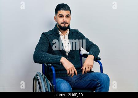 Handsome man with beard sitting on wheelchair puffing cheeks with funny face. mouth inflated with air, crazy expression. Stock Photo