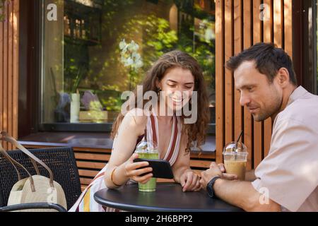 Portrait of happy couple at outdoor cafe looking at smartphone screen while enjoying date in Summer, copy space Stock Photo