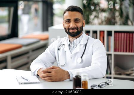 Portrait of a handsome Indian general therapist, in medical uniform and stethoscope, sitting at his desk in the cabinet, in the hospital, looking at the camera, smiling friendly Stock Photo