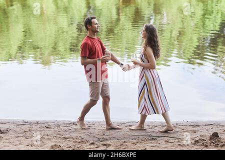 Full length portrait of happy couple holding wine glasses while enjoying romantic date by water outdoors, copy space Stock Photo