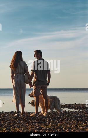 Back lit full length portrait of happy couple with dog together by water at sunset Stock Photo