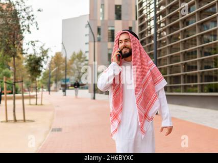 Arab male in traditional clothes smiling and looking away while walking on street and talking on cellphone Stock Photo