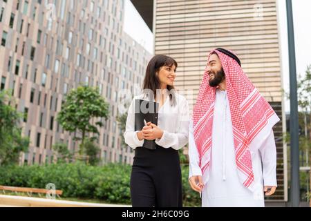 Low angle of happy Arab male smiling and talking with female manager while strolling on street of modern city together Stock Photo
