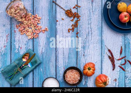 Still life with iridescent metal forks, himalayan salt, colored pasta, tomatoes and peppers on blue wooden background Stock Photo