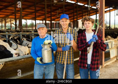 Positive elderly cow breeder with adult son and teenage grandson in cowshed Stock Photo