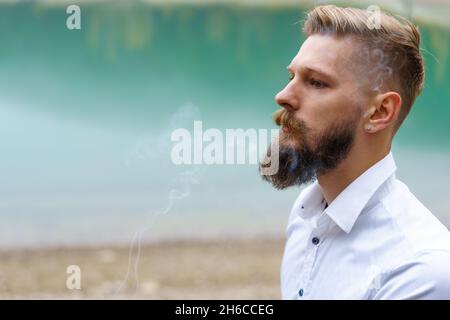 Business caucasian young bearded man in a white shirt smokes a cigarette and blows smoke against the backdrop of a turquoise lake in reflections. Faces solving problems and making choices Stock Photo