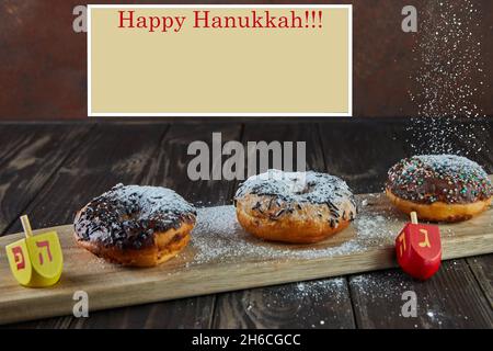 Happy Hanukkah and Hanukkah Sameach - traditional Jewish donuts, spinning tops with pouring powdered sugar on wooden background. Inscription in Hebrew Stock Photo