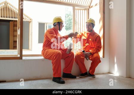 Positive builders in orange overalls sitting on window sill and drinking hot coffee from thermos during break Stock Photo