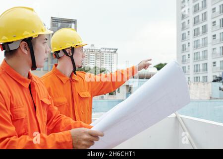 Construction workers standing at site with blueprint in hands and pointing at skyscraper near by Stock Photo