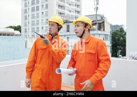 Frowning construction site workers discussing some problem they noticed and talking on walkie-talkie with foreman Stock Photo