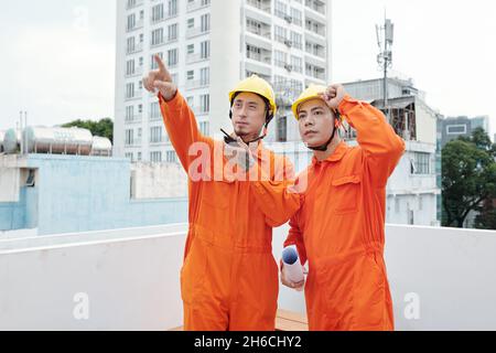 Team of construction site workers in hardhats talking on walkie-talkie and asking colleague to move crane Stock Photo
