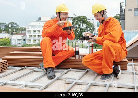Team of smiling builders eating lunch and drinking coffee on rooftop after productive morning at construction site Stock Photo