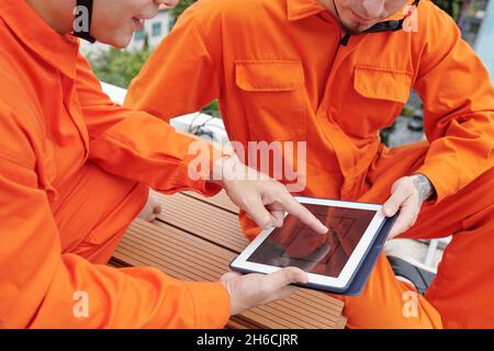 Closeup image of construction workers in bright orange overalls discussing blueprint on digital tablet Stock Photo