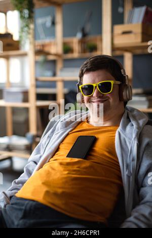Relaxed young man with Down syndrome with sunglasses and headphones sitting indoors at school Stock Photo