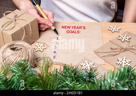 New Year's goals - A hand holding a pen and writing down plans for the new year on a table with gifts, snowflakes and fir branches with balls. Stock Photo