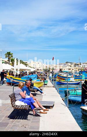 Couple sitting on a bench along the waterfront with fishing boats and pavement cafes to the rear, Marsaxlokk, Malta, Europe. Stock Photo