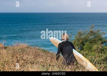 An eager male surfer carrying his surfboard walking down a coastal footpath to the sea in Cornwall. Stock Photo